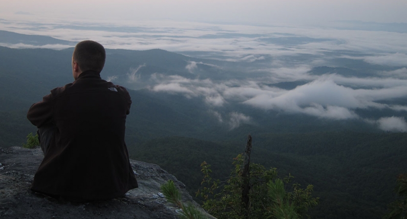 A person sits on a rocky overlook, looking out over the vast forested mountain area below. 
